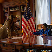 Two women talking in a formal office with wooden bookshelves, a large desk, and an American flag; one is seated and gesturing while the other stands leaning forward. Warm daylight enters through tall windows with sheer curtains.