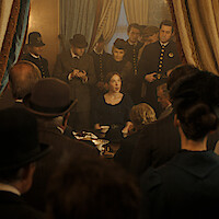 A woman sits at a table surrounded by people in period clothing, with police officers and officials watching her in an ornate room with curtains, suggesting an intense or official meeting scene.