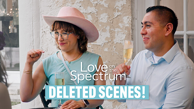 Two people sitting indoors, smiling and holding drinks, with a rustic wall behind them. Text overlay reads "Love on the Spectrum DELETED SCENES!" suggesting this is from a show or video bonus content.