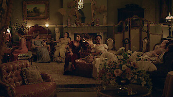 Women in elegant period dresses sit and lounge in a richly decorated parlor with vintage furniture, ornate decor, flowers, and a lit fireplace, creating a warm, historical atmosphere.