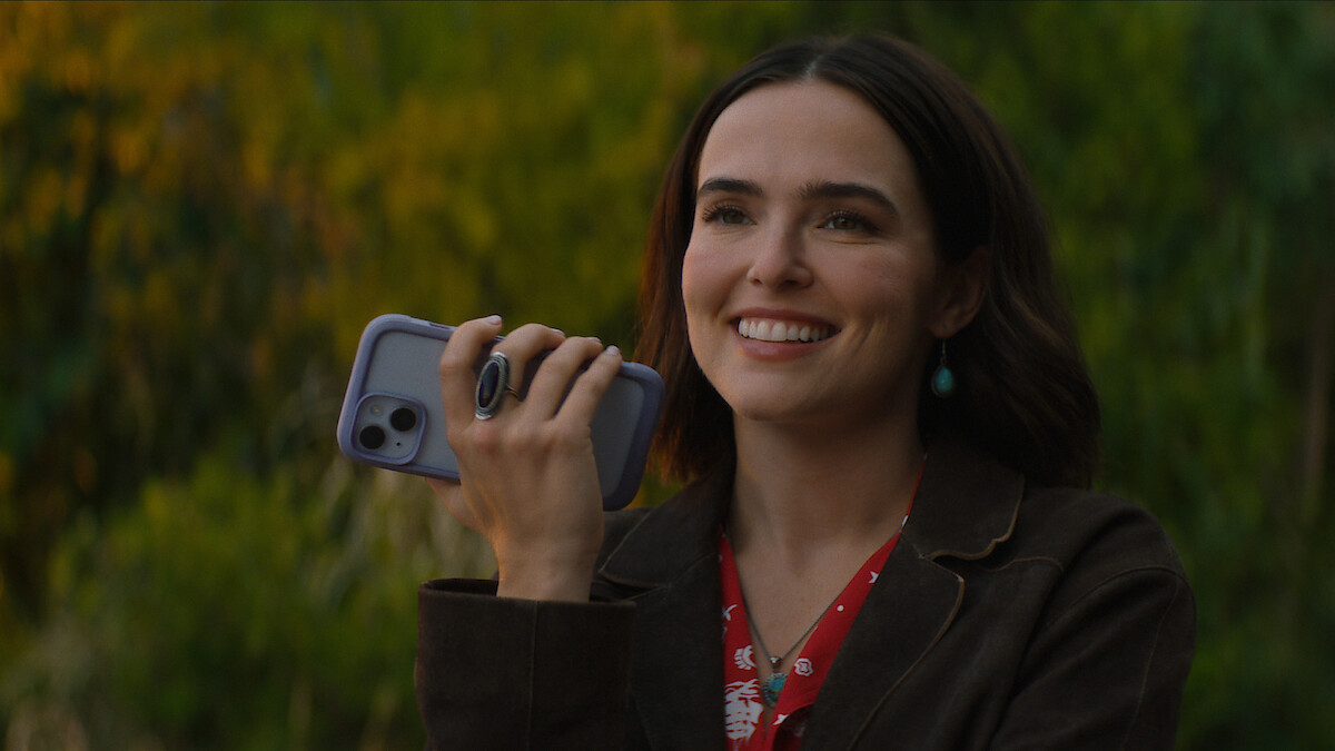 A woman smiling outdoors, holding a smartphone in her hand, wearing a brown jacket and red floral blouse, with green blurred bushes and trees in the background on a sunny day.
