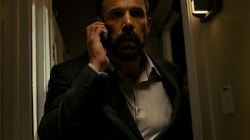 A man in a dark suit talking on a phone walks through a dimly lit hallway, looking serious and focused, with warm lighting on the walls and a tense atmosphere.