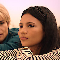 Young woman with long dark hair and striped shirt outdoors, resting her chin on her arms, with a man in a blue shirt beside her, both gazing thoughtfully into the distance in soft natural light.