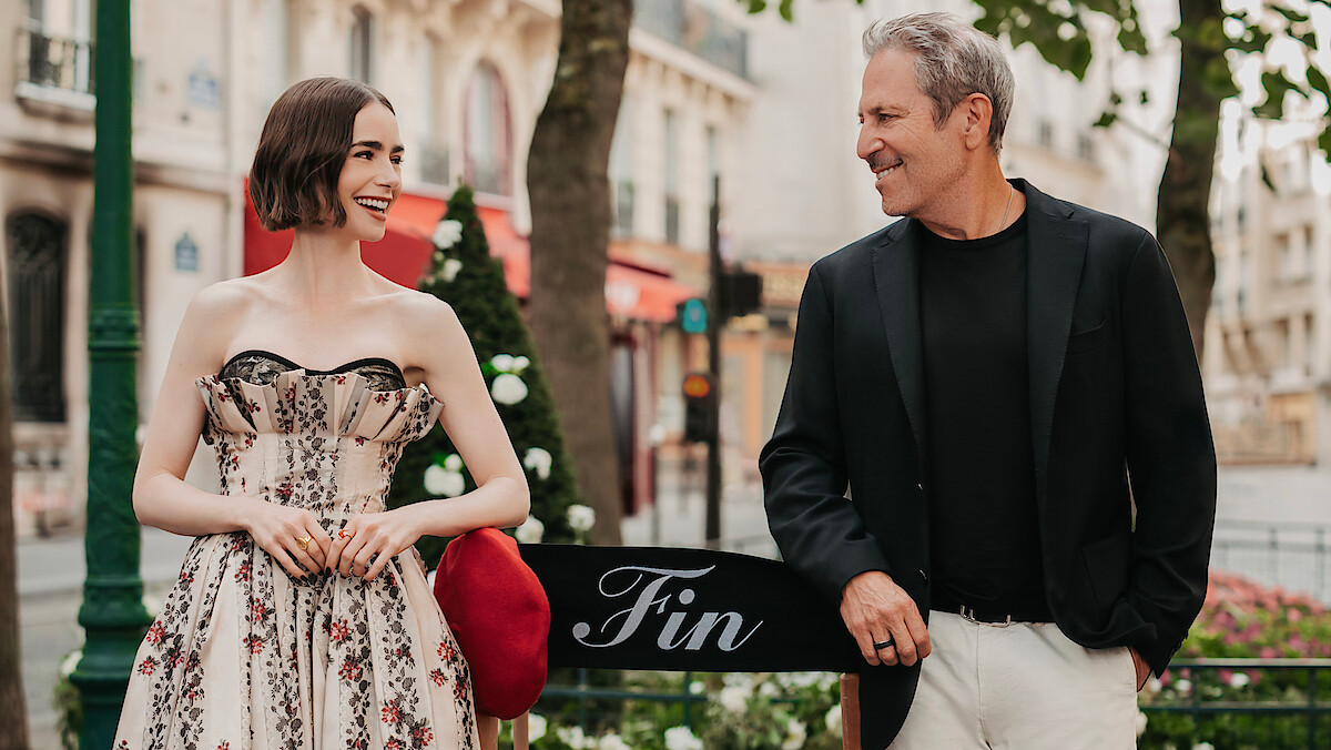 A young woman in a floral dress and a man in a black blazer smile at each other while standing outdoors by a bench labeled "Fin," with trees and classic European buildings in the background.