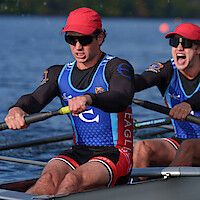 Two male rowers in sunglasses and matching athletic gear intensely rowing a boat on a lake during a race, with blue water, blurred buoys, and trees in the background, expressing focus and teamwork.