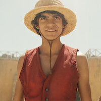 Young person smiling, wearing a straw hat and red vest, standing outdoors on a sunny day with blurred buildings in the background.