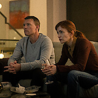 Two people sit on a couch in a dimly lit living room, looking intently at something offscreen. They have takeout containers and drinks on the table, suggesting a casual night at home. Warm lighting creates a cozy and focused atmosphere.