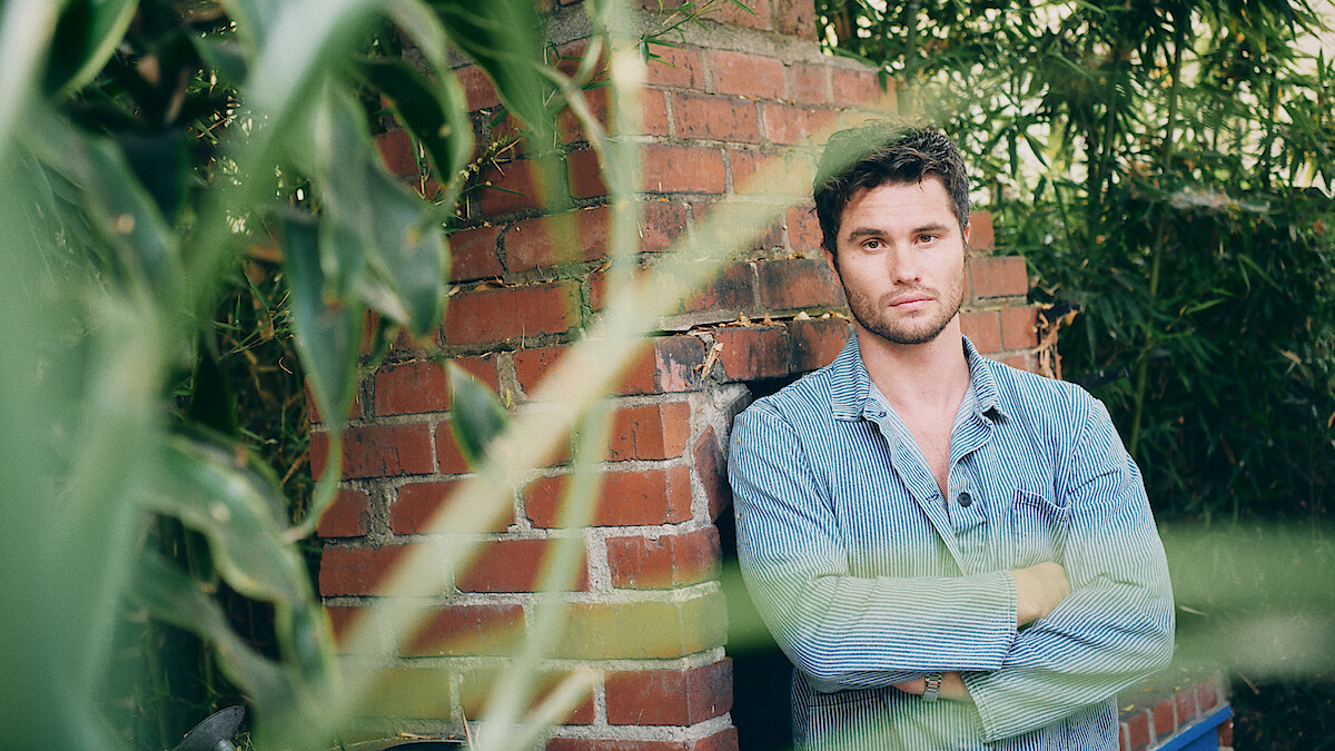 A man in a light denim shirt stands with arms crossed in front of a brick wall outdoors, surrounded by green plants and bamboo. The setting is natural and relaxed, with leaves partially obscuring the foreground.
