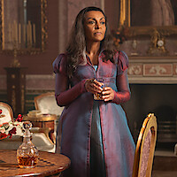 Woman in formal period costume stands holding a glass in an ornate room with classic furniture, a fireplace, floral decor, and a crystal decanter on a wooden table, suggesting a historical, elegant setting.