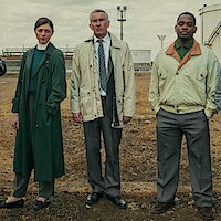 Three people in formal clothing stand outdoors on dry, brown ground near industrial tanks and metal structures, under a cloudy sky. The setting appears to be an industrial or factory facility.