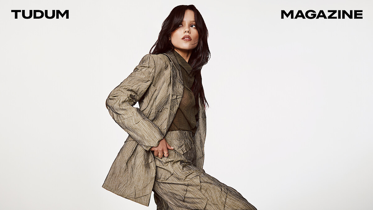 Jenna Ortega in a taupe textured suit with sheer blouse looks to the side, gaze locked on something in the distance with her hair down.