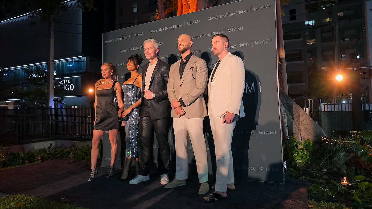 Five well-dressed people posing together on an outdoor evening event stage, city buildings and greenery in the background, creating a formal and celebratory mood.
