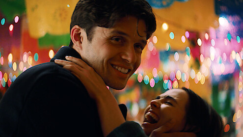 A smiling man embraces a laughing woman at a festive indoor party decorated with colorful lights and banners, creating a joyful, lively atmosphere.