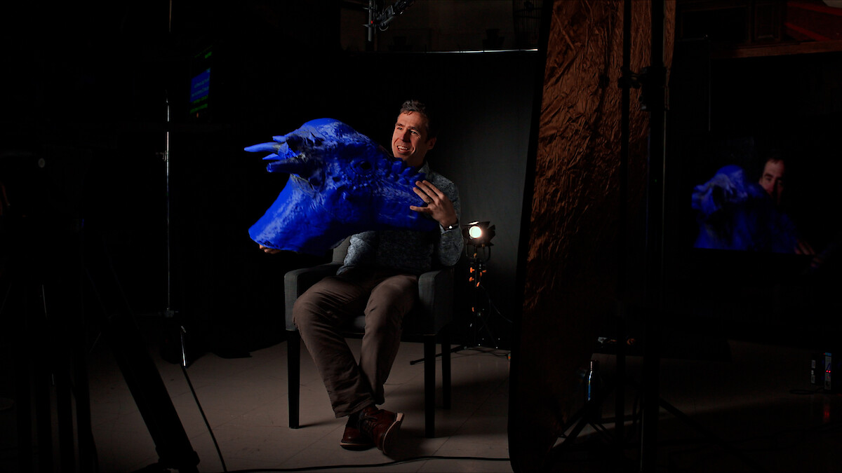 A man holds a prop dinosaur head as he's being interviewed. 