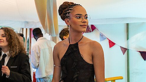 Yasmin Finny in a black dress with braided hair and glasses stands at a festive indoor event with bunting and other guests in formal attire, creating a celebratory atmosphere.