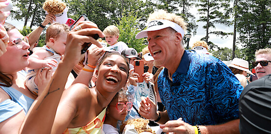 Man in blue shirt and visor smiling and interacting with excited fans outdoors at a daytime event; people taking selfies, trees and bright daylight in the background.