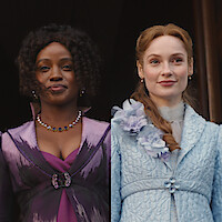 Two women in elegant period costumes, one in purple with a jeweled necklace and one in light blue with floral details, standing side by side against a dark background.