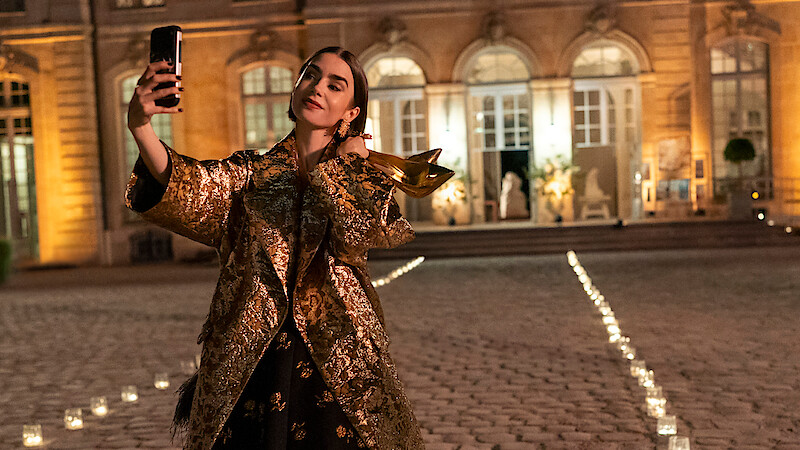 Woman in a gold coat taking a selfie outside a grand, illuminated building at night, with candlelit pathways on a cobblestone courtyard.