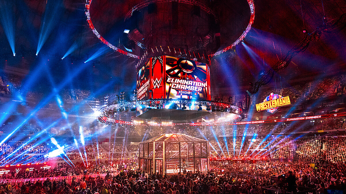 WWE Elimination Chamber wrestling event in a large arena, filled with a cheering crowd, colorful lights, a cage structure in the ring, and giant display screens with event logos and a WrestleMania sign in the background.