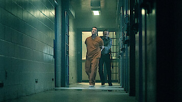A man in an orange prison jumpsuit is being escorted by a guard down a dimly lit hallway in a jail or prison, with barred cells and concrete walls visible.