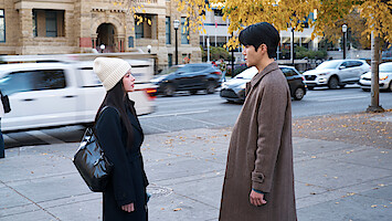 Two people stand facing each other on a city sidewalk lined with autumn trees, wearing coats, with blurred cars and historic buildings in the background.