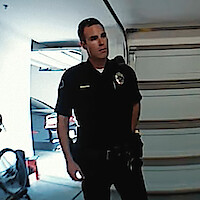 A police officer standing in a garage with a closed garage door behind him, dressed in uniform, hands behind his back, with a car and some equipment visible in the background.