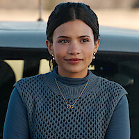 Nikki Rodriguez as Jackie wears a blue sweater and stands by a car in 'My Life with the Walter Boys' Season 2.
