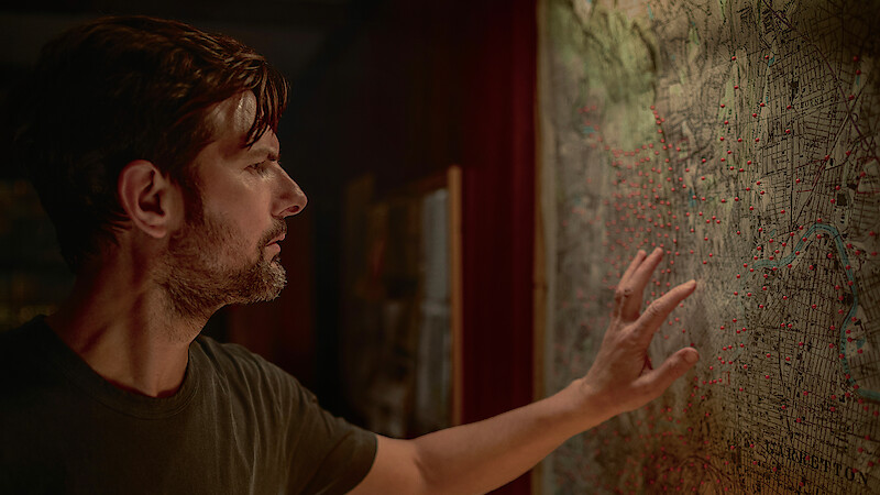 Man in dimly lit room closely examining a large, detailed map on the wall, touching the surface with his hand as he studies numerous red dots marked on the map.