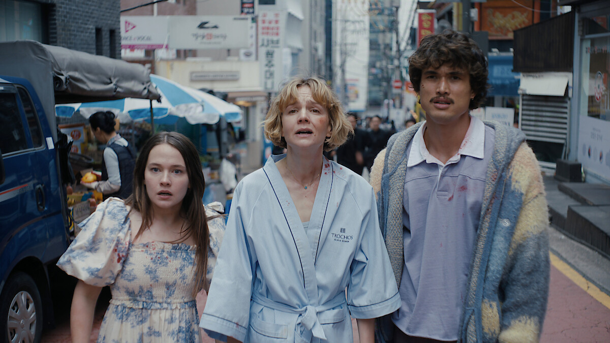 Cailee Spaeny as Ashley Miller, Carey Mulligan as Lindsay Crane-Martin, and Charles Melton as Austin Davis in casual clothing stand on a busy city street, looking upward with surprised expressions, surrounded by vendors, shops, and tall buildings.