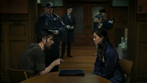 A man and a woman sit across from each other at a table in a dimly lit police interrogation room while officers and investigators work in the background.