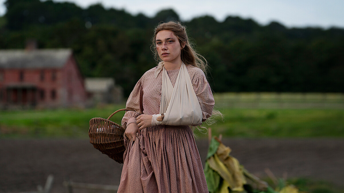 Woman in a long, old-fashioned dress with an arm in a sling walks outdoors near a field, holding a wicker basket. A rustic farmhouse and green trees are visible in the background, suggesting a rural historical setting.