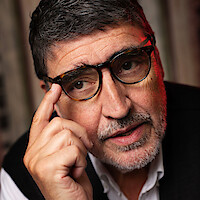 Alfred Molina with his hand raised to his glasses wearing a sweater vest and white button up shirt.