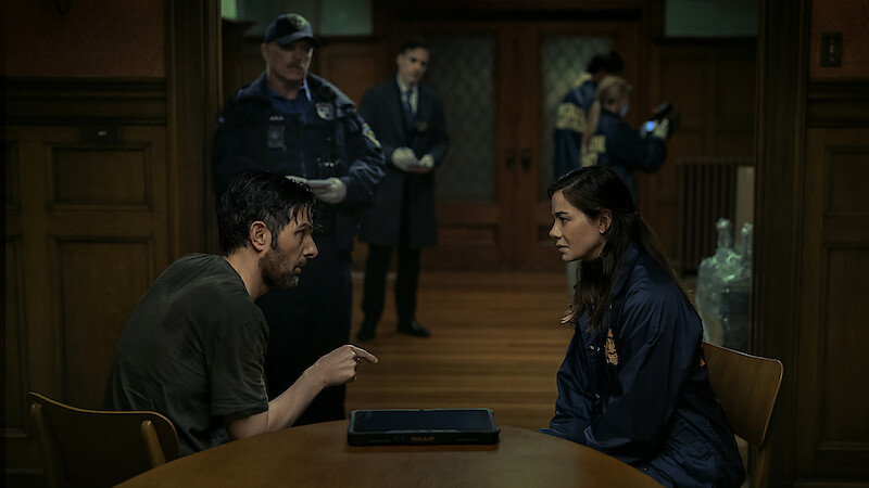 A man and a woman sit across from each other at a table in a dimly lit police interrogation room while officers and investigators work in the background.