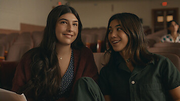 Two young women sit closely together in an auditorium, smiling and talking, with rows of empty seats behind them, creating a warm and friendly atmosphere.