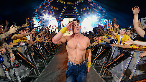 Shirtless wrestler saluting and walking down a ramp, surrounded by cheering fans at a brightly lit arena event, with colorful lights and stage setup in the background.