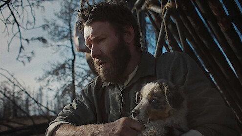 A bearded man sits outdoors in a rustic, wooden shelter, holding a small dog in his arms. The environment looks cold and barren, evoking a somber, contemplative mood as sunlight filters through the sparse trees.