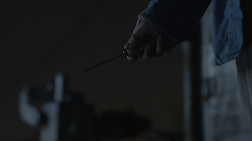 A person holding a knife in their right hand at night, wearing a dark blue jacket. The background is dimly lit, creating a suspenseful or tense atmosphere.