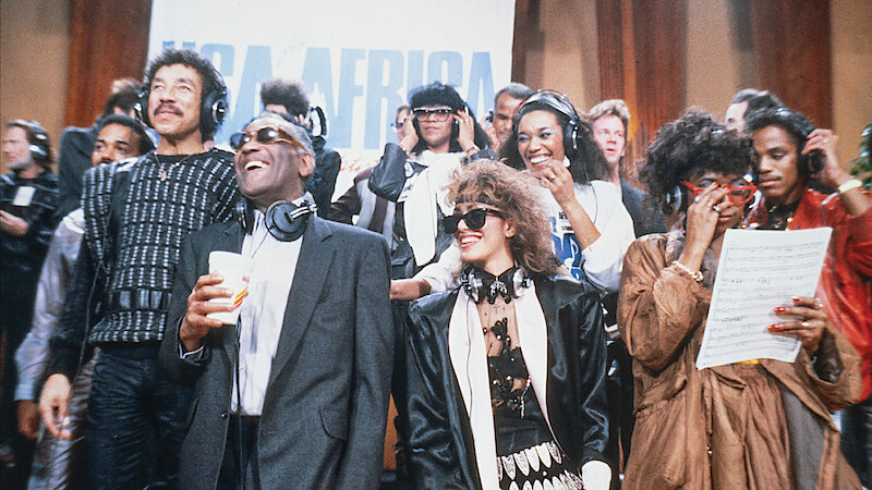 A diverse group of musicians in a recording studio, wearing headphones, singing and smiling together in front of a USA for Africa sign, with music sheets and microphones in a lively, collaborative atmosphere.