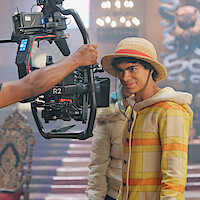 Iñaki Godoy as Monkey D. Luffy, in a yellow plaid costume and straw hat, stands on a decorated film set, facing a professional camera rig held by a crew member, with cinematic lighting and ornate background elements visible.