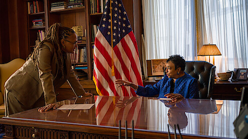 Two women in an office, one standing and one seated behind a polished desk, discuss something. An American flag, bookshelves, and a lamp are in the background, creating a formal, professional setting.