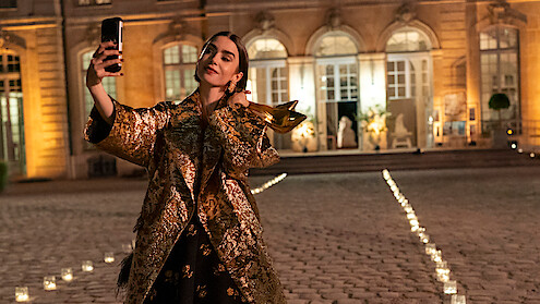 Woman in a gold coat taking a selfie outside a grand, illuminated building at night, with candlelit pathways on a cobblestone courtyard.