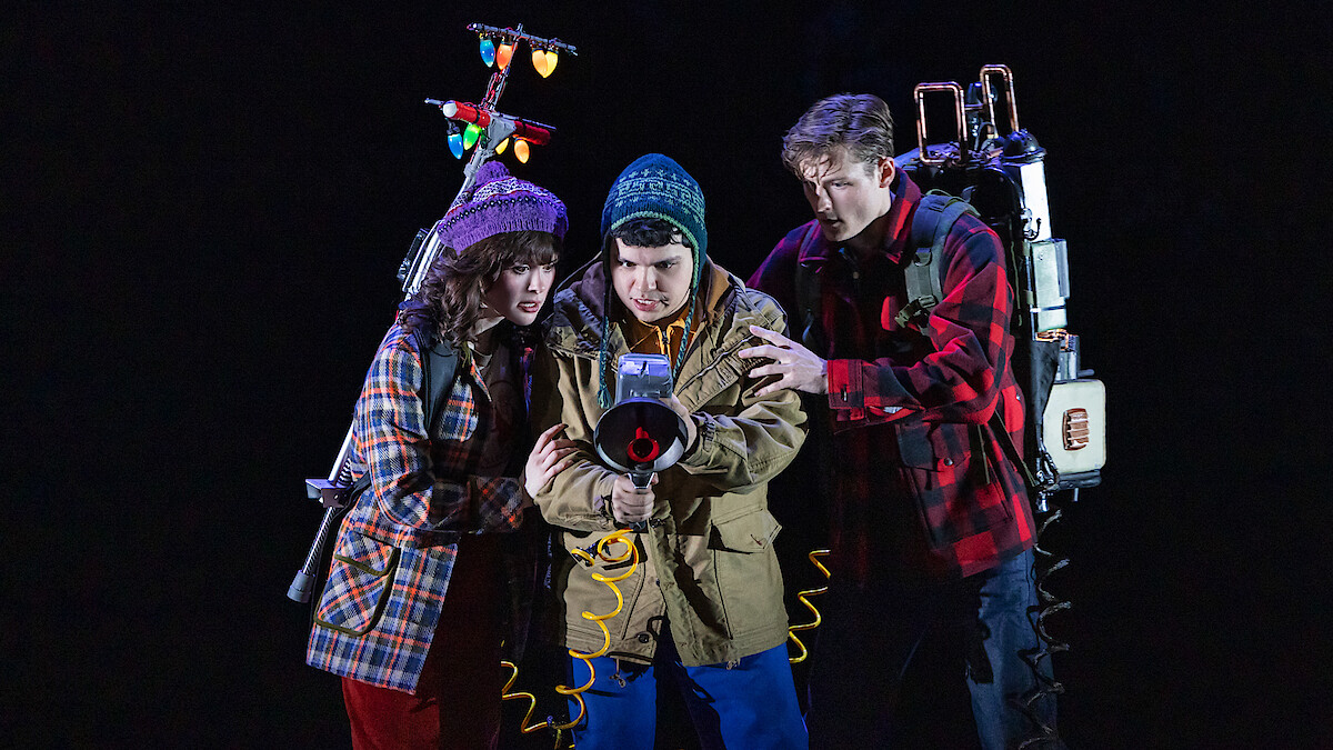 Three young people in winter clothes, holding ghost-busting equipment with glowing lights, stand closely together in a dark, dramatic setting, appearing focused and ready for action.