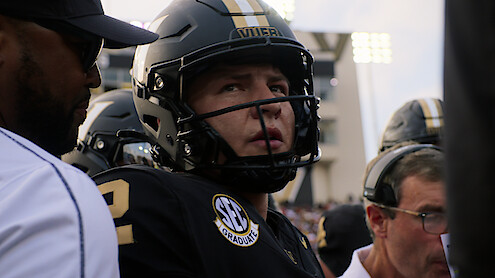 Football player in black uniform and helmet stands with coaches on the sideline during a game, stadium lights in background, focused and intense atmosphere.