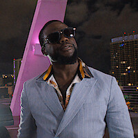Man in sunglasses and a light blue suit stands confidently on a rooftop at night, with city lights and modern buildings in the background, illuminated by purple lighting.