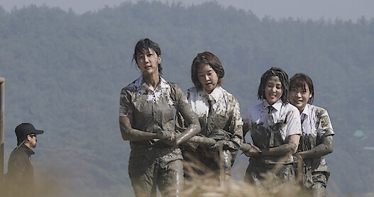 Four women in white short-sleeved button-up shirts, their outfits covered in mud, walk together while carrying a heavy object.