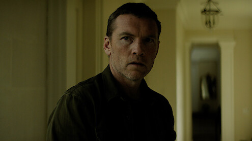 A serious man stands indoors in a dimly lit hallway with pale yellow walls and columns, looking slightly off-camera. The background features a corridor and a hanging light fixture.