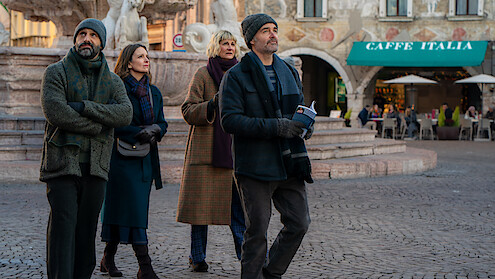 Four adults in winter clothing walk together in a European plaza near a fountain, with a café and people dining outside in the background, suggesting a casual sightseeing outing in an urban historic setting.