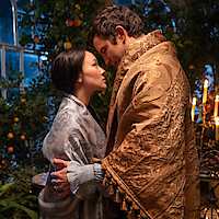 A man and woman stand close, wrapped in ornate shawls, gazing into each other's eyes in a candlelit room filled with greenery and oranges, creating a romantic and intimate atmosphere.