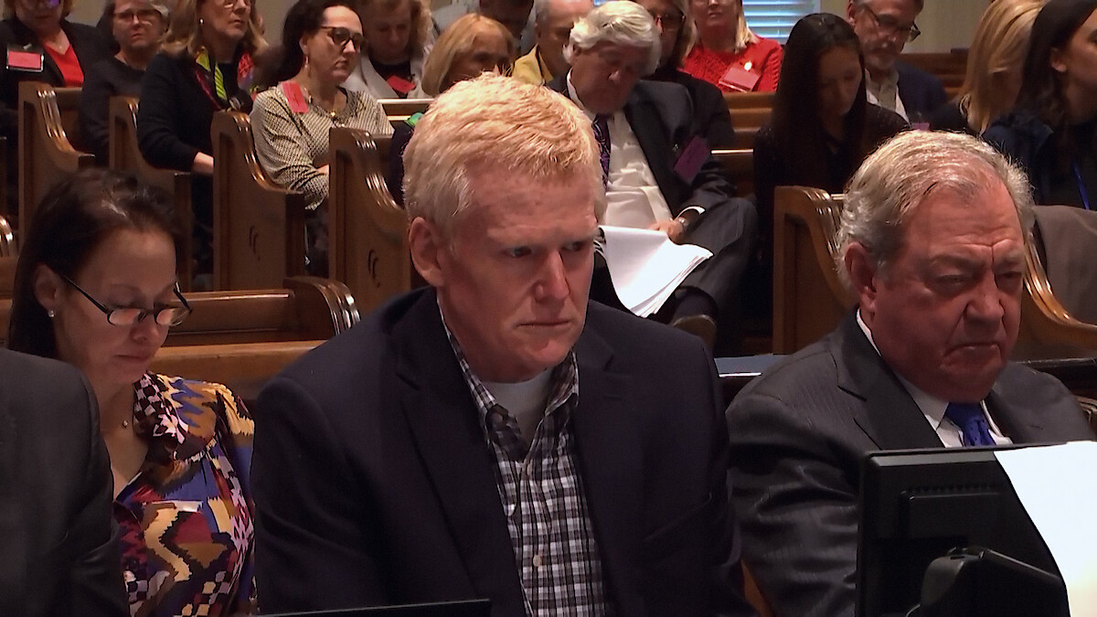 Three people sit at a courtroom table, looking serious. They are surrounded by other people seated in wooden pews, suggesting a legal proceeding in progress. The atmosphere is formal and attentive.