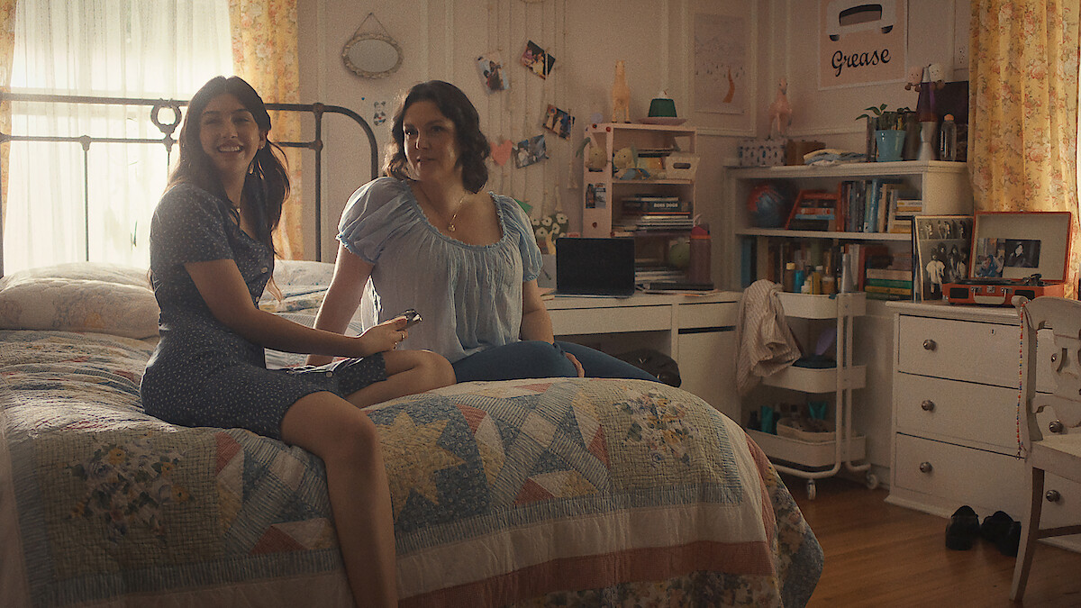 Two women sitting on a bed in a cozy, sunlit bedroom with bookshelves, photos, and personal items in the background, creating a warm and inviting atmosphere.